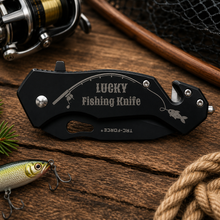 Load image into Gallery viewer, A black folding fishing knife engraved with 'LUCKY Fishing Knife' and a fishing line graphic, displayed on a rustic wooden surface with a fishing reel, lure, rope, and net in the background.
